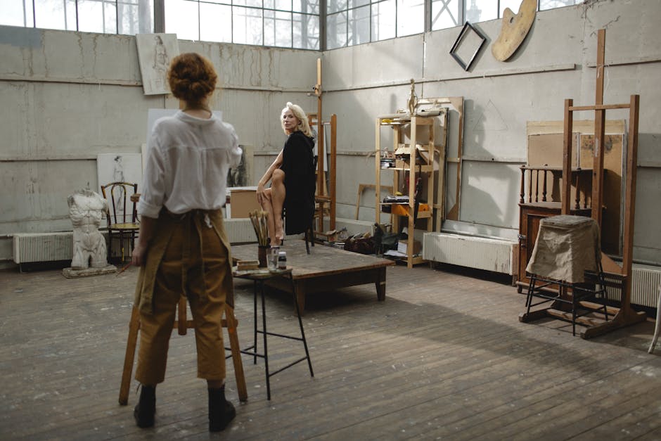 Two women engaged in artistic work inside a bright, spacious studio filled with art supplies and easels.